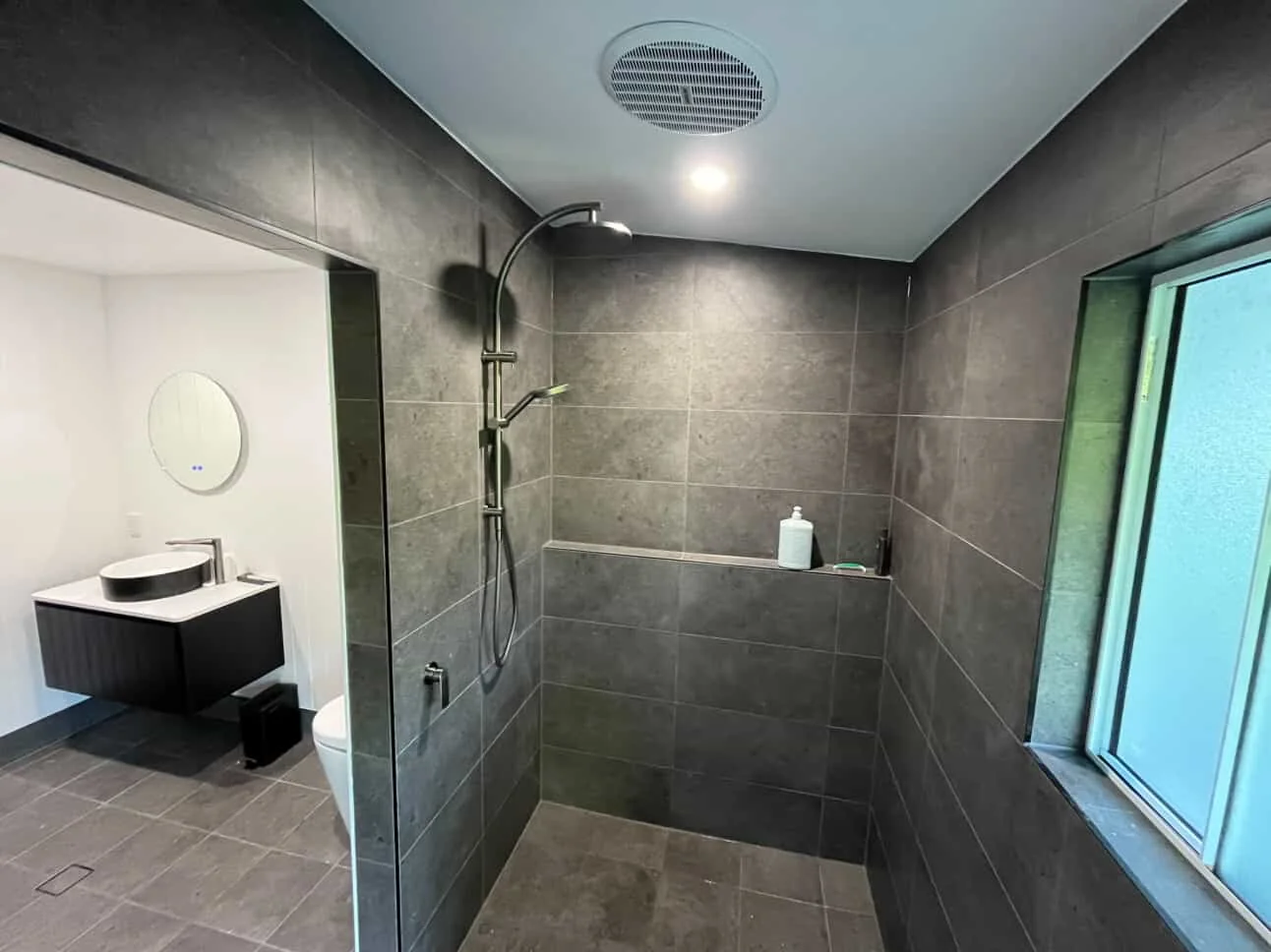 Dark grey tiled walk-in shower with glass screen, black shower fittings, recessed shelf and a window, beside a wall-hung vanity
