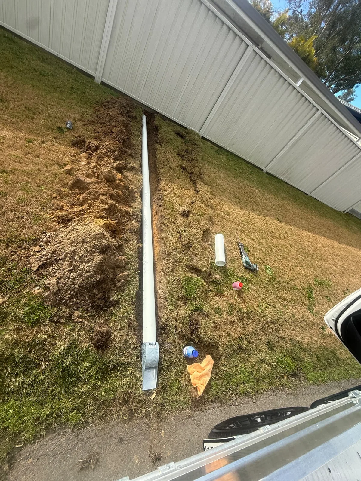 White pipe running through a narrow soil trench beside a metal fence